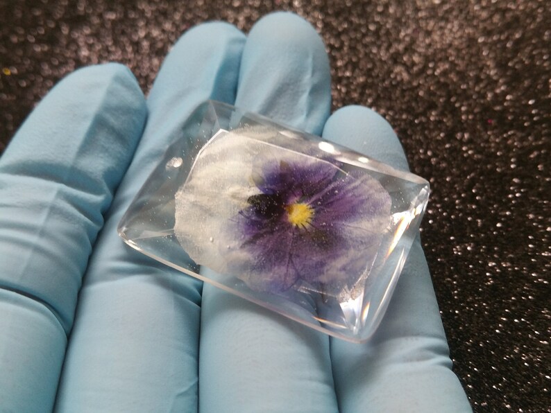 K&ouml;nnte beinhalten: Ein klarer, rechteckiger Harzanh&auml;nger mit einer gepressten lila-wei&szlig;en Stiefm&uuml;tterchenblume. Die gelbe Mitte der Blume ist sichtbar. Der Anh&auml;nger wird in einer behandschuhten Hand gehalten.