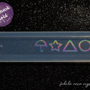 Pode incluir: Molde de silicone transparente para fazer marcadores de página. O molde tem quatro desenhos: um guarda-chuva, uma estrela, um triângulo e um círculo. O molde está sobre um fundo preto com glitter.