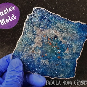 May include: A blue and silver coaster mold with a silver edge. The coaster has a geode-like appearance with a mix of blue, silver, and copper-coloured accents. The text "Coaster Mold" is in a purple circle.
