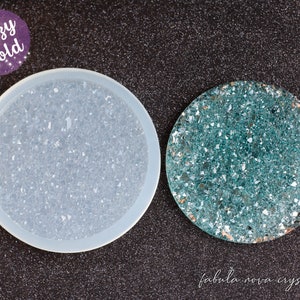 May include: A round, light blue silicone mold filled with sparkling crystals next to a finished teal coaster with the text "fabula nova crystallis". The mold is labeled "Druzy Mold" in a purple circle.