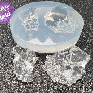 May include: A clear silicone mould with two crystal-shaped cavities, and two finished crystal castings. The crystals are multifaceted and clear, with a sparkling appearance. The text "Druzy Mold" is in a purple circle. The text "fabula nova crystallis" is at the bottom.