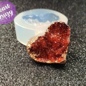 May include: A red heart-shaped druzy with gold accents. The druzy is made of small crystals and has a sparkly finish. The druzy is sitting on a white silicone mould.
