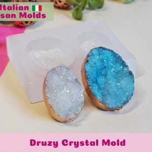 May include: A clear silicone mold with two cavities, next to two druzy crystal pendants. One pendant is light blue, the other is white, both with gold-colored edges. The text "Italian Artisan Molds" and "Druzy Crystal Mold" are visible.
