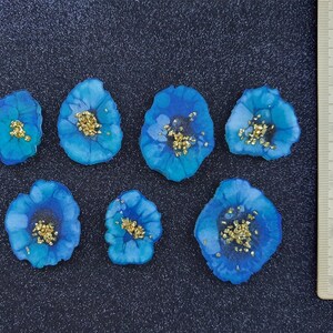 May include: Seven blue resin flower embellishments with gold glitter accents. The flowers are approximately 1 inch in diameter.