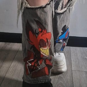 May include: Gray denim jeans with distressed details, featuring graphic illustrations and text. The jeans have ripped knees and a relaxed fit. The text on the jeans reads "THIS FACE WAS MADE FOR RAD" and "THERES A BRA DAY".