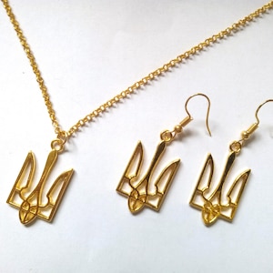 Pode incluir: Colar e brincos de ouro com um design de tridente estilizado. O pingente e os brincos são feitos de metal dourado e têm um design delicado e intrincado.