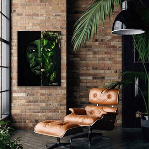 May include: A painting of a person's face with a green and black color scheme. The painting is hanging on a brick wall in a room with a leather armchair and ottoman.