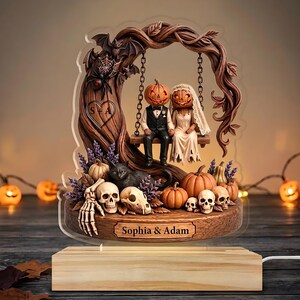 May include: A decorative acrylic lamp featuring a carved tree with a swing holding two pumpkin-headed figures in wedding attire. The base displays the text Sophia & Adam, surrounded by skulls, a black cat, pumpkins, and a heart with S+A.