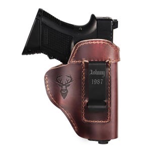 May include: A black handgun in a brown leather holster. The holster has a deer head emblem and the text "Johnny 1987" on the clip. The holster is designed for concealed carry.