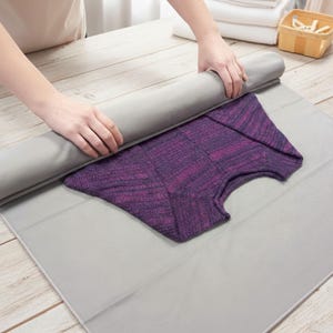 Può includere: Una persona arrotola un tessuto grigio su un maglione viola piegato su una superficie grigia. Il maglione ha un motivo strutturato. La scena è ambientata su un tavolo di legno chiaro, con una pila di asciugamani bianchi e un cestino sullo sfondo.