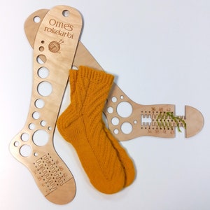 Könnte beinhalten: Zwei hölzerne Sockenstricknadel-Messgeräte mit Löchern zum Messen verschiedener Größen. Die Messgeräte sind mit dem Text "Omes rokdarbi" beschriftet und haben Zahlen, die verschiedene Größen angeben. Eine gelbe gestrickte Socke liegt auf den Messgeräten.