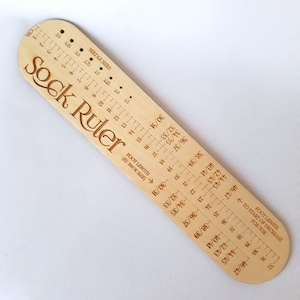 Könnte beinhalten: Ein hölzernes Sockenlineal mit eingravierten Markierungen für Nadelstärken und Fußlänge in EU-Schuhgrößen. Der Text "Sock Ruler" ist deutlich sichtbar. Das Lineal ist hellbraun.