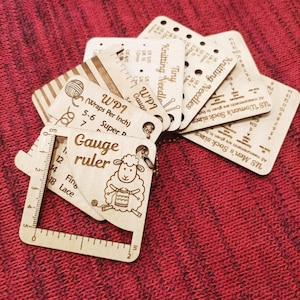 May include: A set of light-coloured wooden knitting gauge tools on a metal ring. The tools feature engraved text including Gauge ruler, WPI, and Knitting Needles, along with a sheep illustration. The ruler shows measurements in inches and centimetres.
