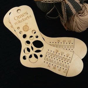 Könnte beinhalten: Ein hölzerner Sockenmaßstab mit Löchern zum Messen verschiedener Größen. Der Maßstab ist mit "Omes rokdarbi" beschriftet und hat eine kleine Illustration einer Wollknäuel und Stricknadeln. Der Maßstab hat auch Markierungen für die Größen 34, 36, 38, 40, 41, 42 und 43.