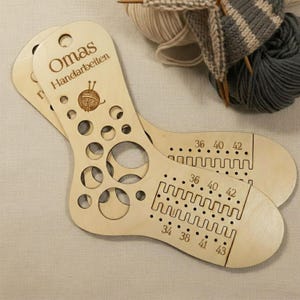 May include: Two wooden sock knitting gauges with circular cutouts and size markings. The text "Omas Handarbeiten" and a yarn ball graphic are etched on the top. Knitting needles and yarn are in the background.