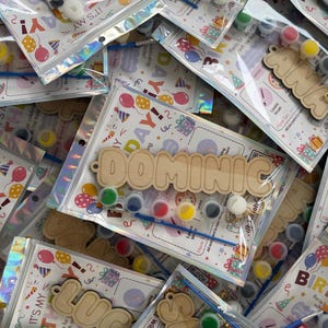 May include: A collection of birthday-themed craft kits in clear plastic bags. Each kit contains a wooden name cutout, a small blue paintbrush, and a strip of six colourful paint pots. The backing card features balloons, gifts, and the text IT'S MY BIRTHDAY.