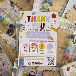 May include: A rectangular thank you card featuring colourful illustrations of school supplies, a rainbow, and an apple. The text reads Thank You For Being Part Of My Class, listing contents including a paint brush, six paint colours, a custom wooden design, and a keyring attachment, with a QR code at the bottom.