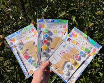 May include: Three birthday-themed paint-your-own wooden character kits in holographic resealable bags. Each kit includes a wooden cutout, a small paintbrush, and six small circular paint pots in various colors. The packaging features the text IT'S MY BIRTHDAY.