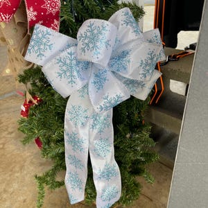 May include: A white satin ribbon bow with a blue snowflake pattern. The bow is tied around a green Christmas tree.
