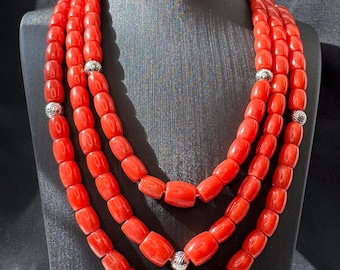 May include: A three-strand necklace featuring vibrant red barrel-shaped beads. The necklace is accented with silver-colored, spherical spacers. The necklace is displayed on a black mannequin.