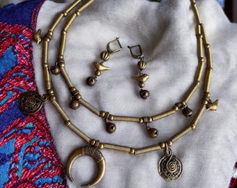May include: A gold necklace with a crescent moon pendant, two small bird charms, and a circular charm with a spiral design. The necklace is made of coiled wire and has small bells hanging from it. Matching earrings with a bird charm and a bell are also included.