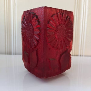 May include: A red glass vase with a square shape and a floral design. The vase has a textured surface and is decorated with raised flowers and leaves.