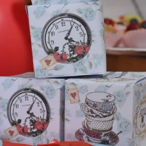 Puede incluir: Tres cajas blancas con dise&ntilde;os de tem&aacute;tica de Alicia en el Pa&iacute;s de las Maravillas. La caja superior tiene una esfera de reloj con rosas y cartas de juego. La caja del medio tiene una esfera de reloj con rosas. La caja inferior tiene una pila de tazas de t&eacute; y platillos con un coraz&oacute;n y el texto "Azul 5 a&ntilde;os".