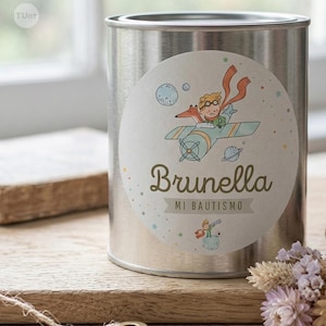Può includere: Un contenitore in metallo argentato con un adesivo rotondo. L'adesivo mostra un'illustrazione a cartoni animati di un ragazzo su un aereo con una volpe, con il nome "Brunella" e il testo "MI BAUTISMO". Lo sfondo include stelle e pianeti.