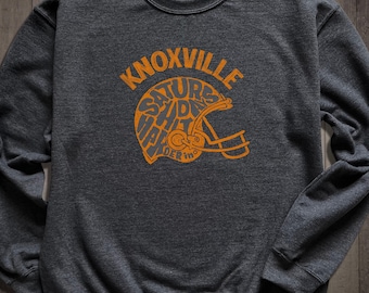 May include: A dark grey sweatshirt with an orange graphic print of a football helmet with the words "KNOXVILLE" and "SATURDAYS IN THE VOLS" inside the helmet.
