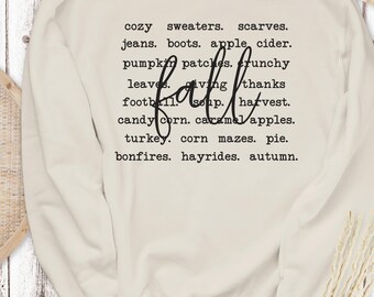 May include: Cream-colored sweatshirt with the word "fall" in a cursive font. The sweatshirt features a list of autumn-themed words in a black, typewriter-style font, including "cozy sweaters," "pumpkin patches," and "bonfires."