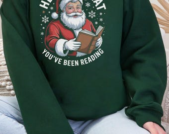Op de afbeelding: Donkergroene sweatshirt met een afbeelding van de Kerstman die een boek leest. De tekst "HE SEES WHAT YOU'VE BEEN READING" staat boven en onder de illustratie van de Kerstman. Het ontwerp bevat sneeuwvlokken.