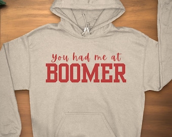 Puede incluir: Una sudadera con capucha gris claro con el texto "you had me at BOOMER" impreso en rojo.