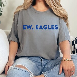 May include: Gray t-shirt with the blue text "EW, EAGLES". The shirt is a crew neck style and is paired with light blue ripped jeans. The model is wearing a gold bracelet and rings.