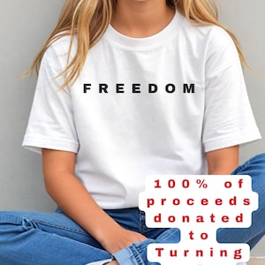 Könnte beinhalten: Weißes T-Shirt mit dem Wort "FREEDOM" in schwarzen Blockbuchstaben. Das Shirt wird mit blauen Jeans und weißen Turnschuhen getragen. Der Text auf dem Bild besagt, dass 100 % des Erlöses an Turning Point USA gespendet werden.