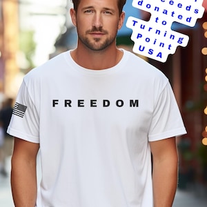 May include: White t-shirt with the word "FREEDOM" printed in black block letters. An American flag is on the left sleeve. The image also includes text that says "100% of proceeds donated to Turning Point USA."