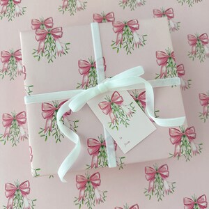 May include: A wrapped gift on pink wrapping paper with a repeating pattern of pink bows and mistletoe. The gift is tied with a white ribbon and a matching gift tag that says "to from".