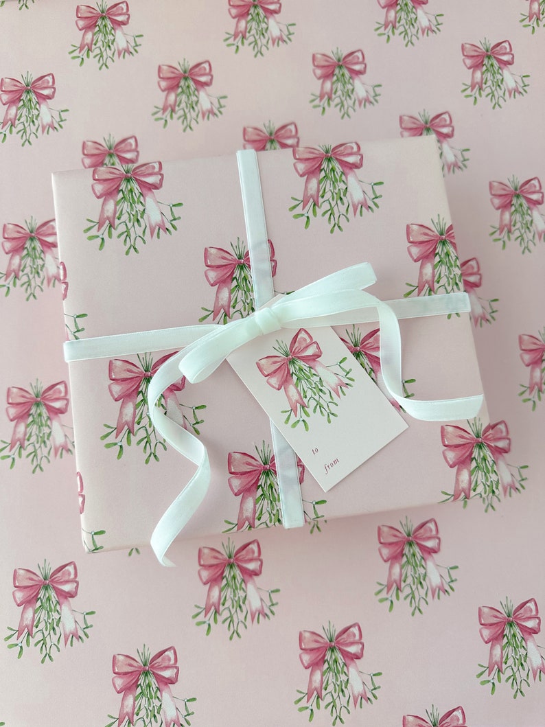 May include: A wrapped gift on pink wrapping paper with a repeating pattern of pink bows and mistletoe. The gift is tied with a white ribbon and a matching gift tag that says "to from".