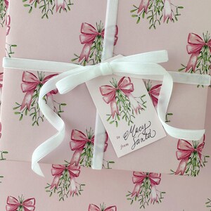 May include: Pink wrapping paper with a repeating pattern of watercolor bows and mistletoe. A white ribbon wraps around the package, and a gift tag reads "to Macy from Santa."