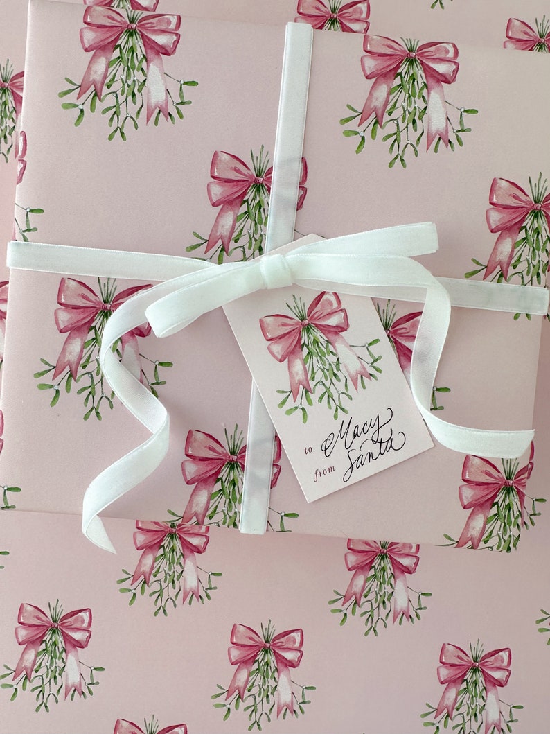 May include: Pink wrapping paper with a repeating pattern of watercolor bows and mistletoe. A white ribbon wraps around the package, and a gift tag reads "to Macy from Santa."