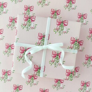 May include: A wrapped gift with a white ribbon on a pink background. The wrapping paper features a repeating pattern of pink bows and green mistletoe. The ribbon is tied in a bow on top of the gift.