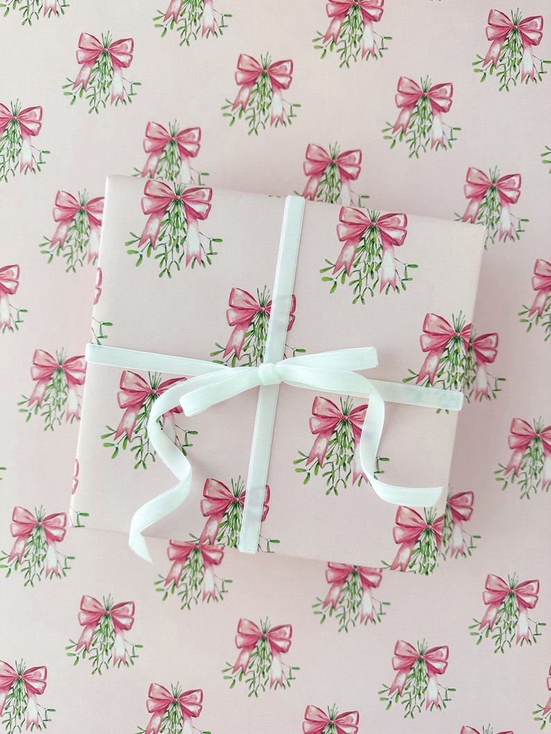 May include: A wrapped gift with a white ribbon on a pink background. The wrapping paper features a repeating pattern of pink bows and green mistletoe. The ribbon is tied in a bow on top of the gift.
