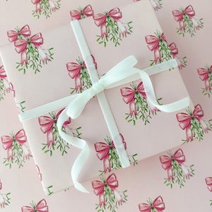 May include: A wrapped gift with a light pink background and a repeating pattern of pink bows and green mistletoe. The gift is tied with a white ribbon. The wrapping paper and ribbon create a festive and elegant presentation.