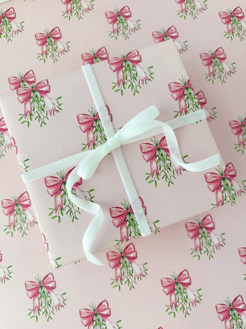 May include: A wrapped gift with a light pink background and a repeating pattern of pink bows and green mistletoe. The gift is tied with a white ribbon. The wrapping paper and ribbon create a festive and elegant presentation.