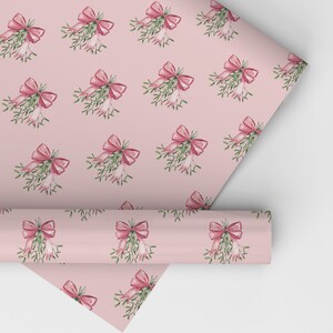 May include: Pink wrapping paper with a repeating pattern of watercolor mistletoe and bows. The bows are a deep pink color, and the mistletoe has green leaves and white berries. A roll of the paper is partially unrolled.