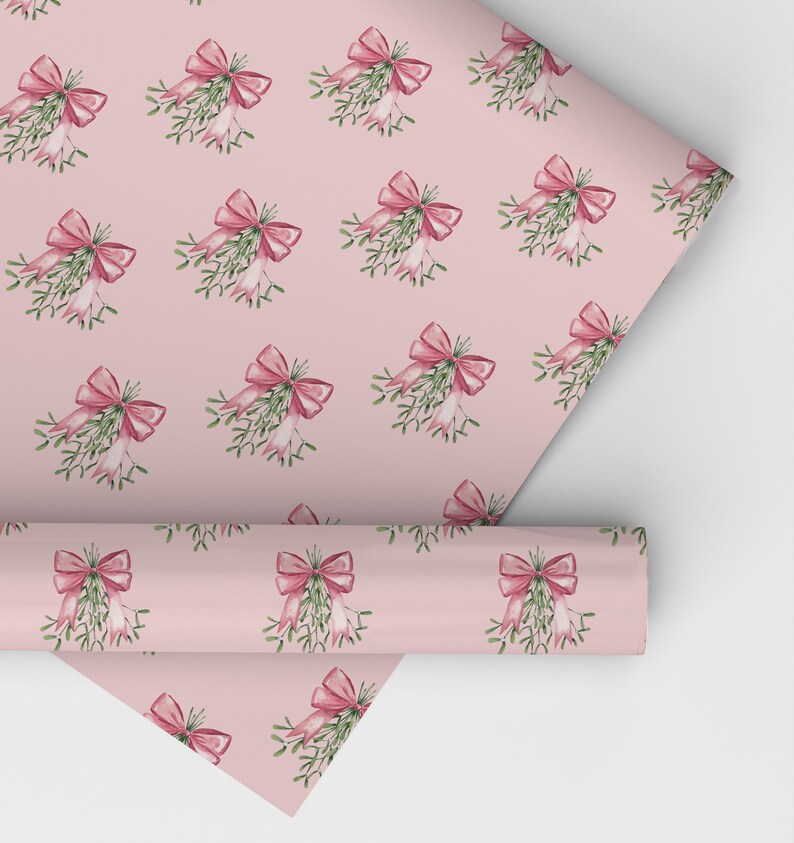 May include: Pink wrapping paper with a repeating pattern of watercolor mistletoe and bows. The bows are a deep pink color, and the mistletoe has green leaves and white berries. A roll of the paper is partially unrolled.