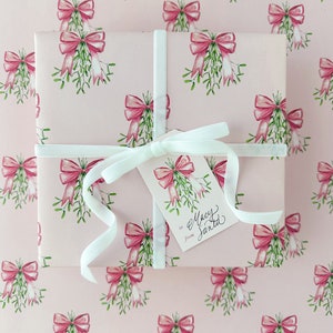 May include: A wrapped gift with pink wrapping paper featuring a pattern of pink bows and green mistletoe. The gift is tied with a white ribbon and a tag that reads "to Macy from Santa."