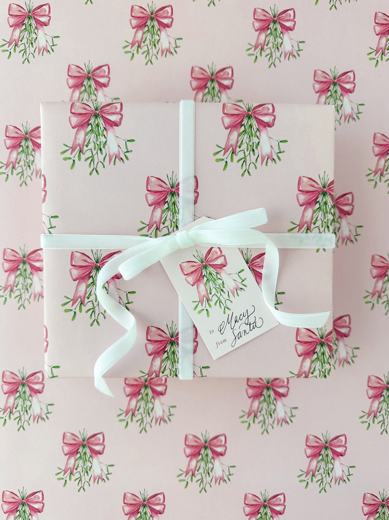 May include: A wrapped gift with pink wrapping paper featuring a pattern of pink bows and green mistletoe. The gift is tied with a white ribbon and a tag that reads "to Macy from Santa."