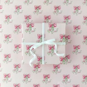 May include: A wrapped gift with a white ribbon on a pink background. The wrapping paper features a repeating pattern of pink bows and green mistletoe. The gift is square and the ribbon is tied in a simple bow.
