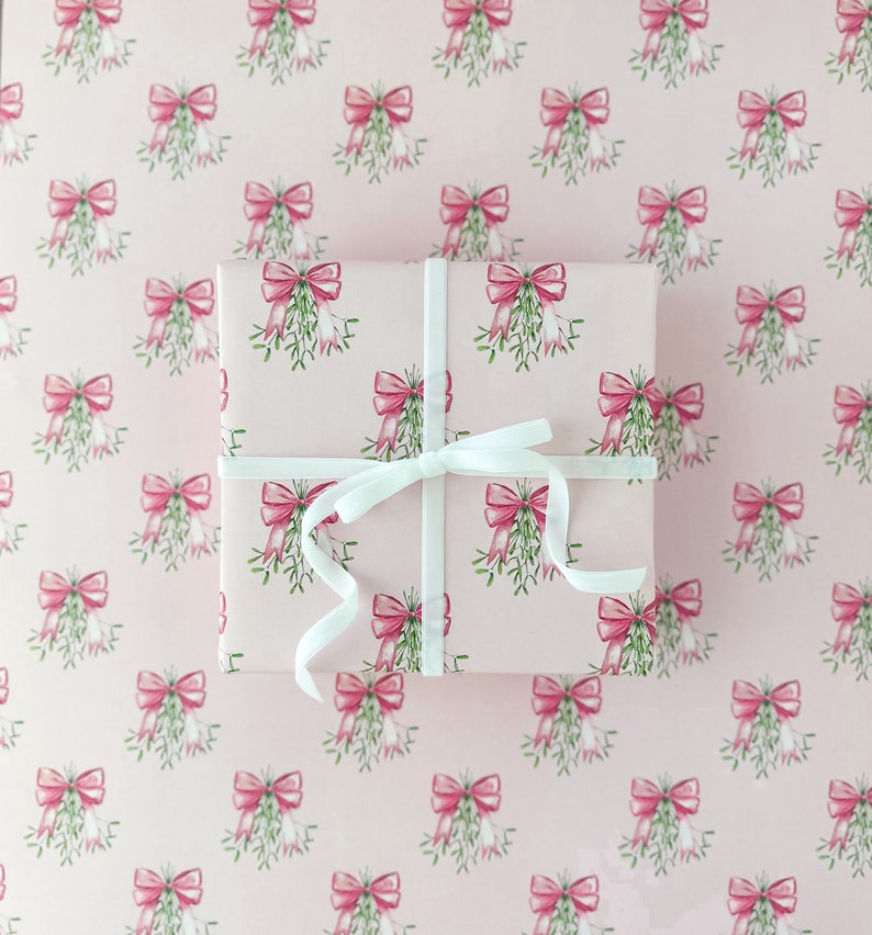 May include: A wrapped gift with a white ribbon on a pink background. The wrapping paper features a repeating pattern of pink bows and green mistletoe. The gift is square and the ribbon is tied in a simple bow.