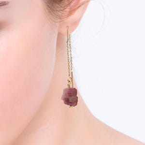 May include: A close-up of a person's ear wearing a gold chain earring with a raw, pink tourmaline gemstone. The earring hangs down, showcasing the natural texture and color of the stone. The background is a soft, neutral tone.